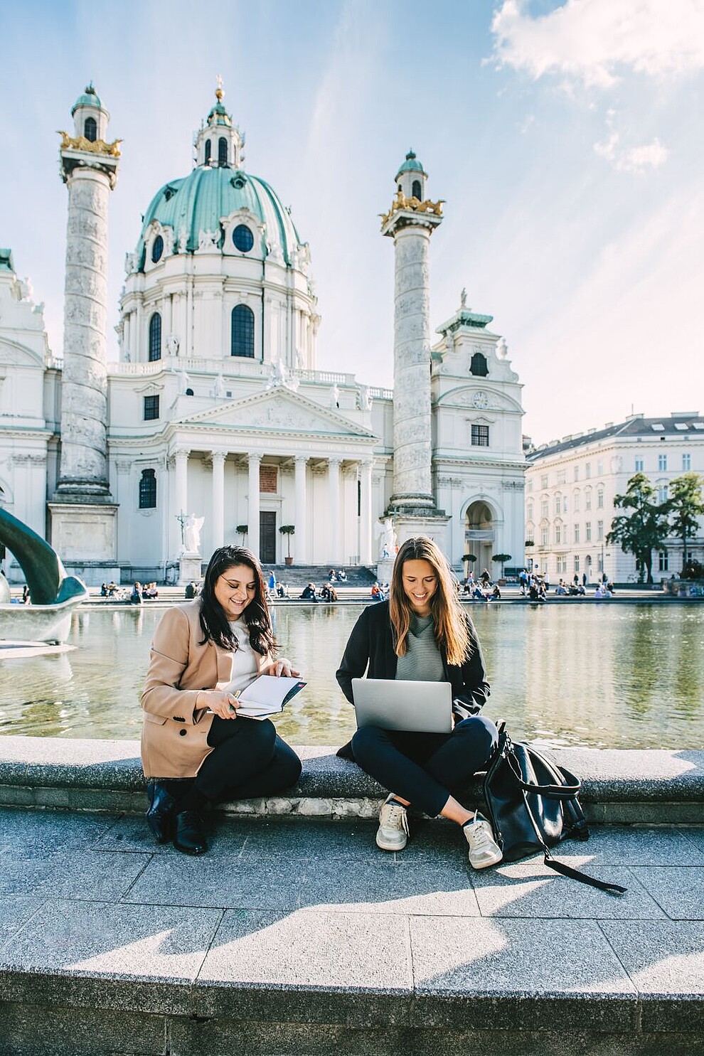 Two women working on the laptop on the edge of a fountain in front of Vienna's Karlskirche.