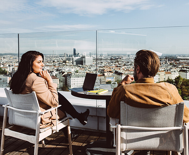 International innovation hub Vienna Woman and man sitting and speaking on a rooftop Vienna cityscape in front of them